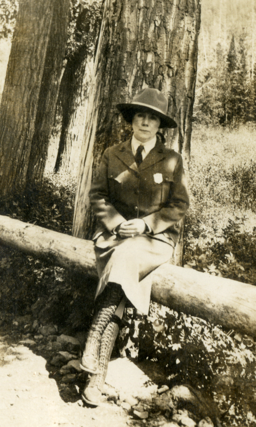 Ranger Irene Wisdom Irene Wisdom sits on a log posing in her NPS uniform skirt, shirt, tie, jacket, and Stetson hat.