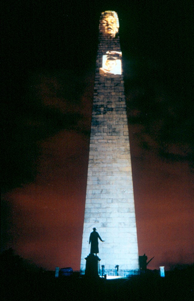 a tall monument in darkness with a projection of a woman's face at the very top