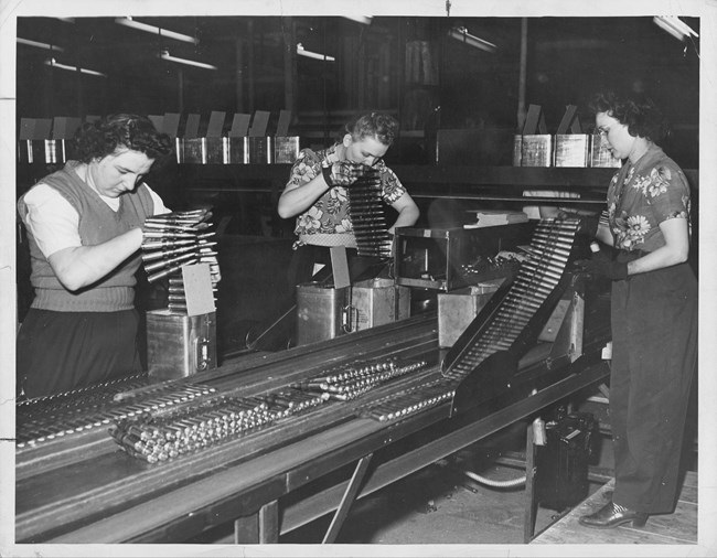 Two women stand on the far side of a production line. They hold long belts of ammunition over metal boxes. A third woman stands on the opposite side of the production line. She monitors the the casings as they emerge from a machine.