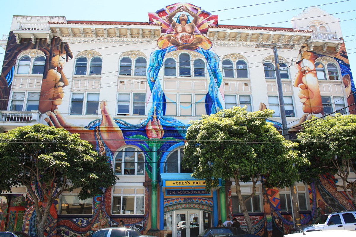 White multi-story building with arched windows covered in mural depicting central figure of seated pregnant woman with butterfly wings over falling water flanked by two faces in profile facing one another