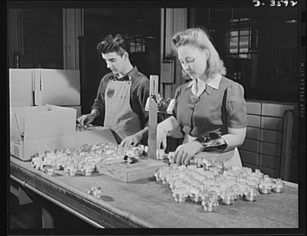 Women work side by side with men to smash the Axis Dark-haired white man and blonde woman test and inspect parts for ammunitions shells. The man wears an apron and the woman wears thick work gloves.