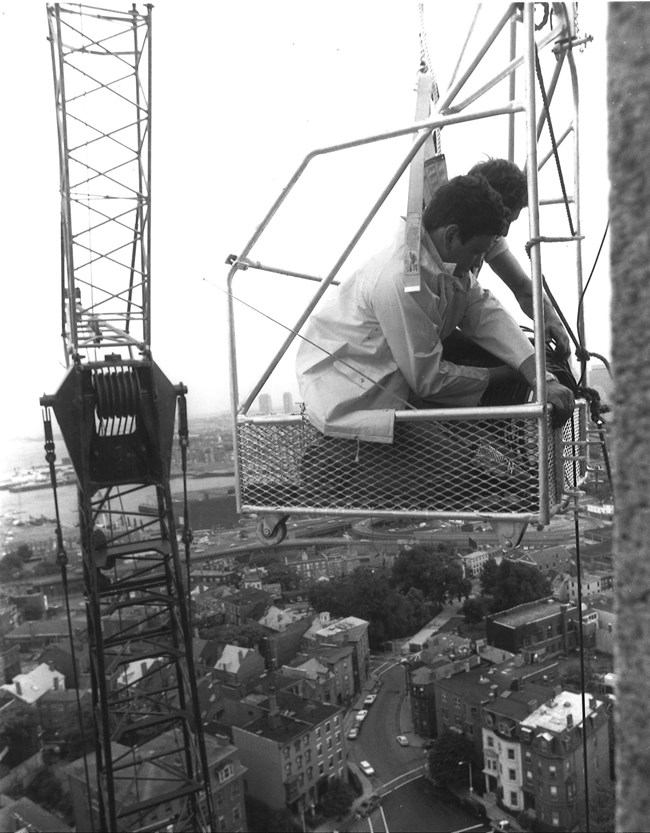 Workers in a small metal carriage that a crane holds up alongside the Bunker Hill Monument.