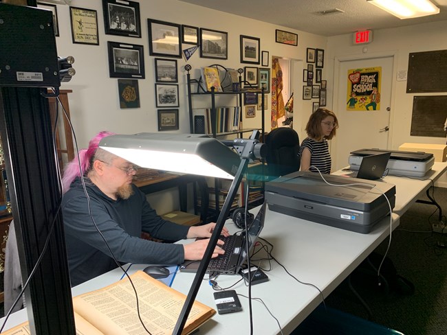 Two people sit at a long table with a set of scanners and laptops