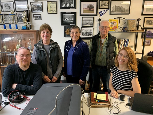 Five people, three standing and two sitting, behind a table with scanners and laptops