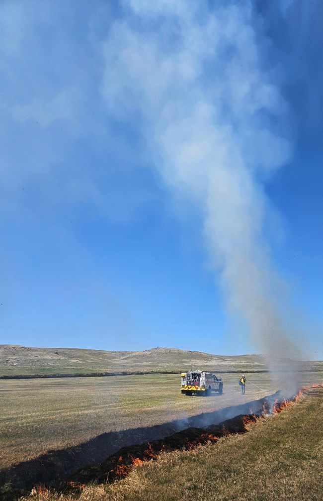 Blackened vegetation and flames from burning grass line a ditch across a field of short green grass. A white fire engine with a wildland firefighter stand along the ditch as smoke rises from the burning grass.