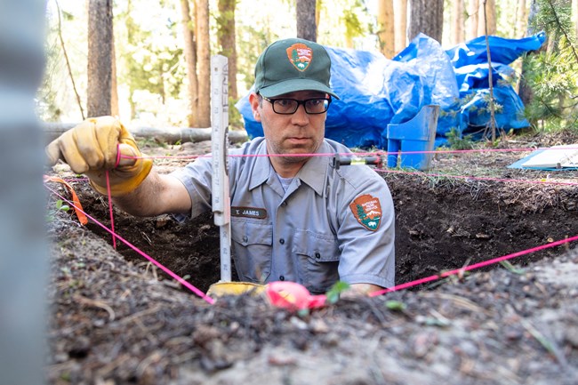 Uniformed National Park Service archaeologist stands in hole and sets up measuring string in Yellowstone National Park.