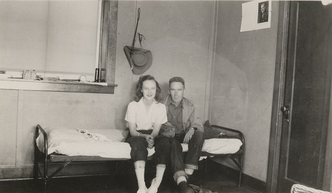 Nancy Bowers sits with a man on a cot in a dingy room below a window. A photo of a solider hangs above her on the wall.