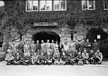 A large group of men and women pose together in three rows in front of a vine covered stone building with a wooden sign for a Museum shown.