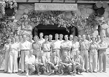 A group of men and women stand in front of a museum entrance. the first row of people are all men and are kneeling on the ground.