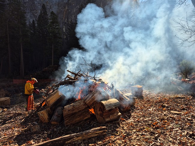 Person standing next to pile of burning logs.