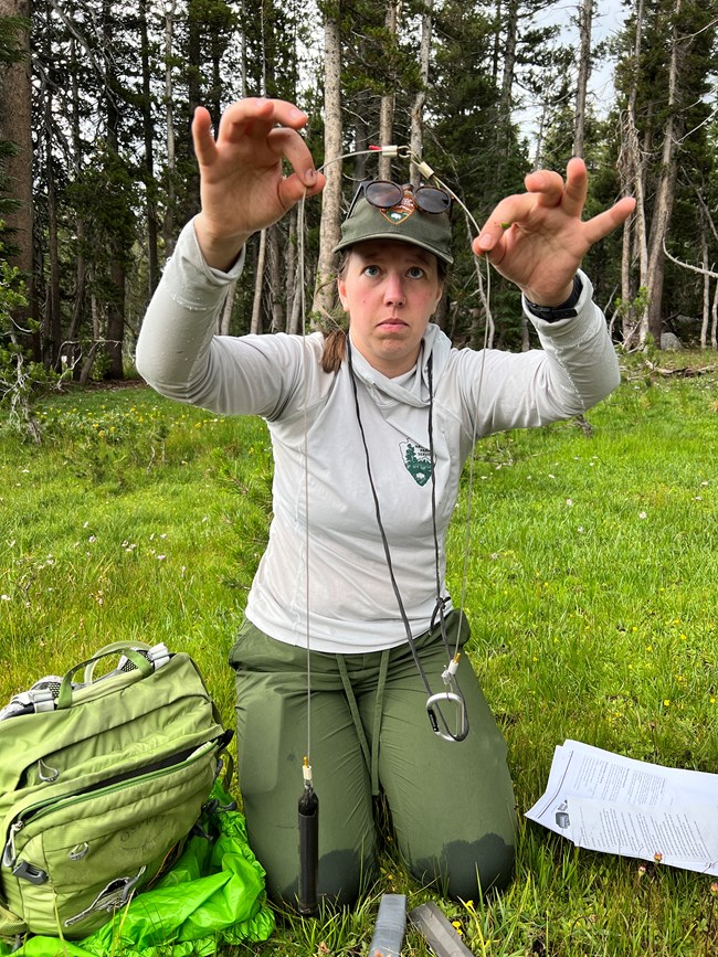 Woman in NPS field uniform kneels on the ground and focuses her attention on a data logger she has removed from a wetland well.