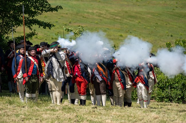 soldiers in revolutionary war uniforms firing muskets shoulder to shoulder