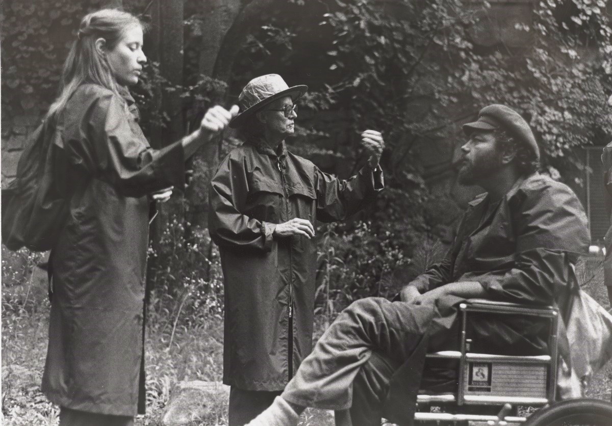 A man and two women outdoors. The man is in a wheelchair. One woman is using sign language to interpret to the man what the other woman is saying.