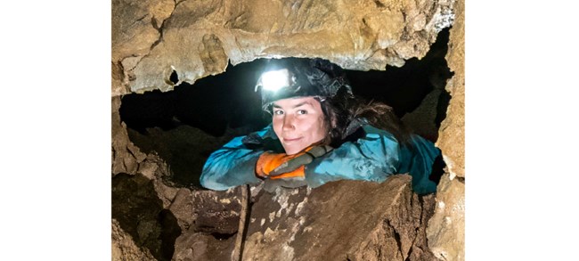 Sierra Heimel Heimel wearing gloves and a headlamp, lying on her stomach, resting her chin on her hands, and smiling down through a cave opening.
