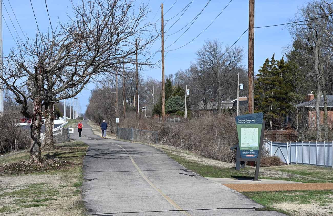 Two people walking on a paved concrete trail, sign that reads "Gravois Greenway Grant's Trail."