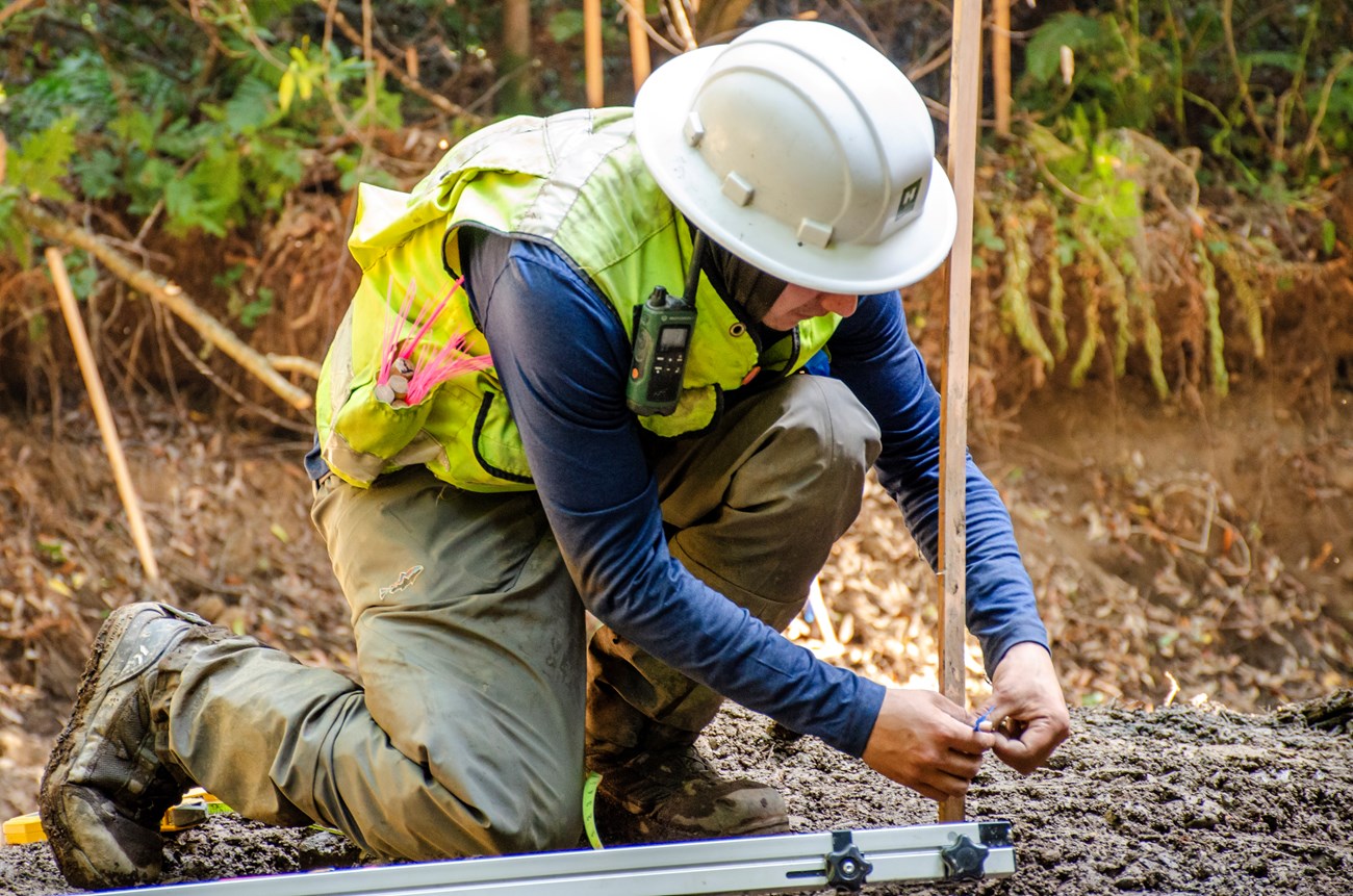 Jeffrey Perez at work Jeffrey in a hard hat and safety vest down on the ground on one knee tying blue flagging tape around the bottom of a wooden stake.