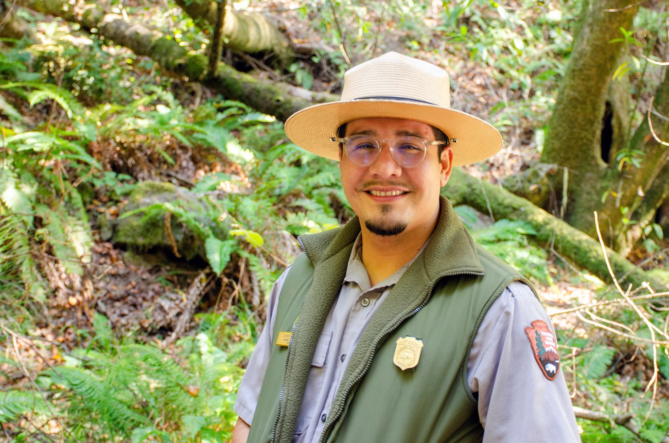 Andres, in glasses and his National Park Service flat hat and uniform, smiles aganst a backdrop of a fern-covered hillside.