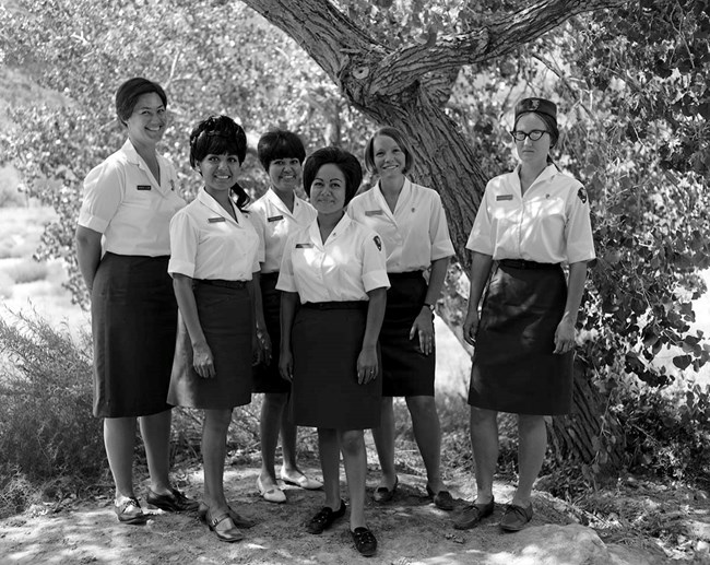 Zion 9557_1969 Uniform Six women all wearing white blouses, dark colored skirts, and oxford style shoes stand together in the shade of a tree.