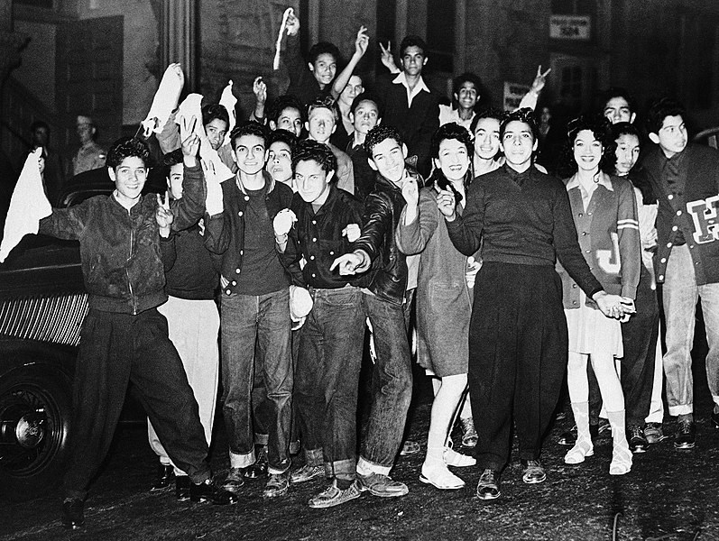 Smiling young Zoot Suit rioters wearing letter sweaters, jeans, and wide leg pants make V signs