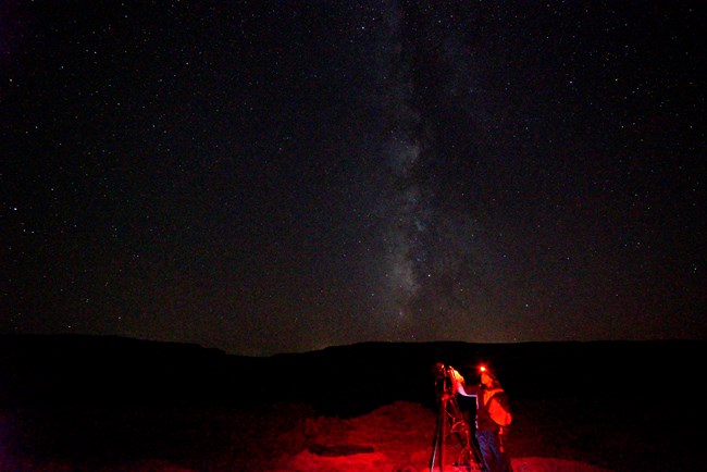 photo of a night sky and mountains in the background with a young woman and a camera on a tripod in a red glow from a headlamp on Li-Wei's head