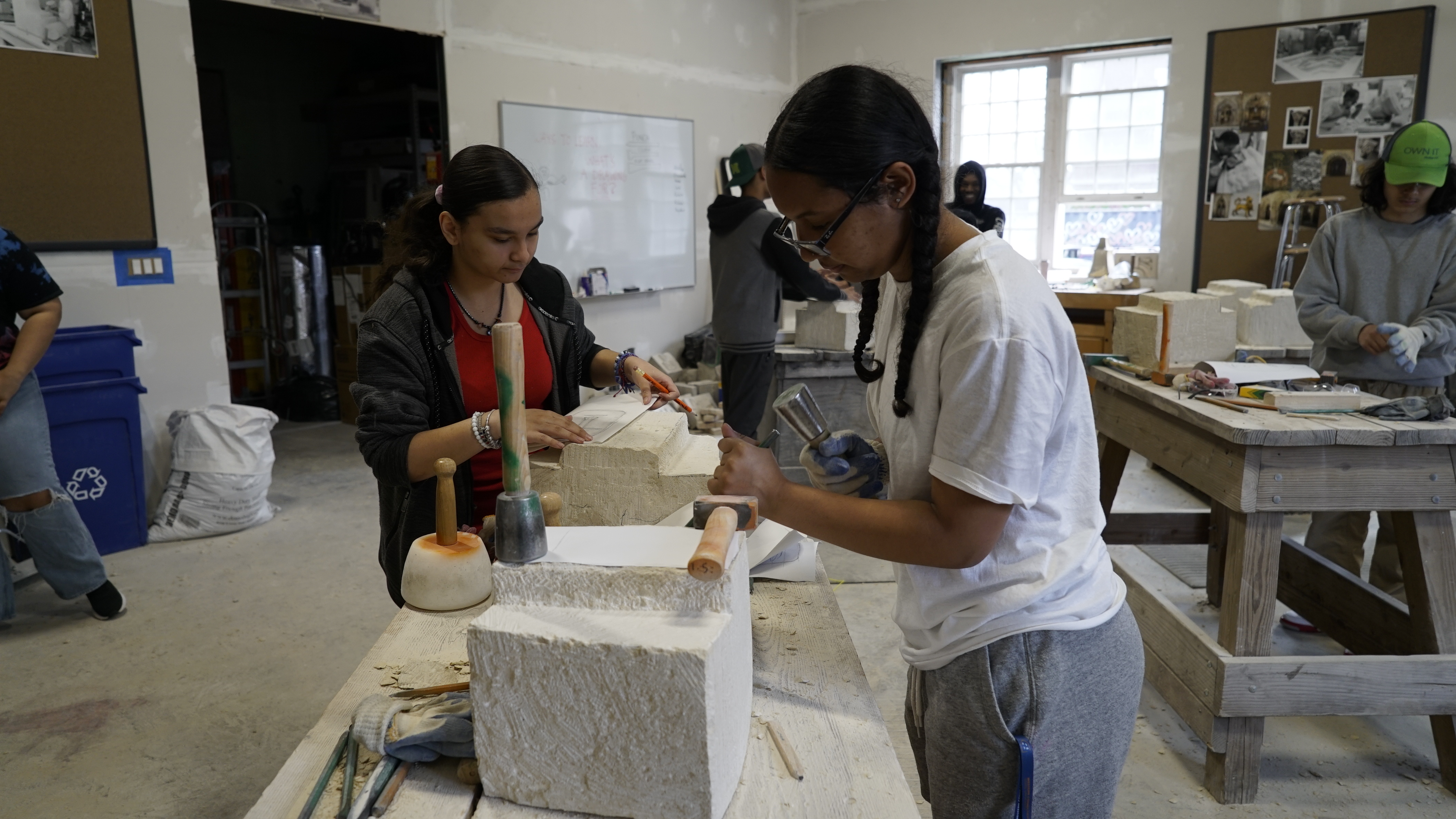 Two young women stand at a work table and carve stone