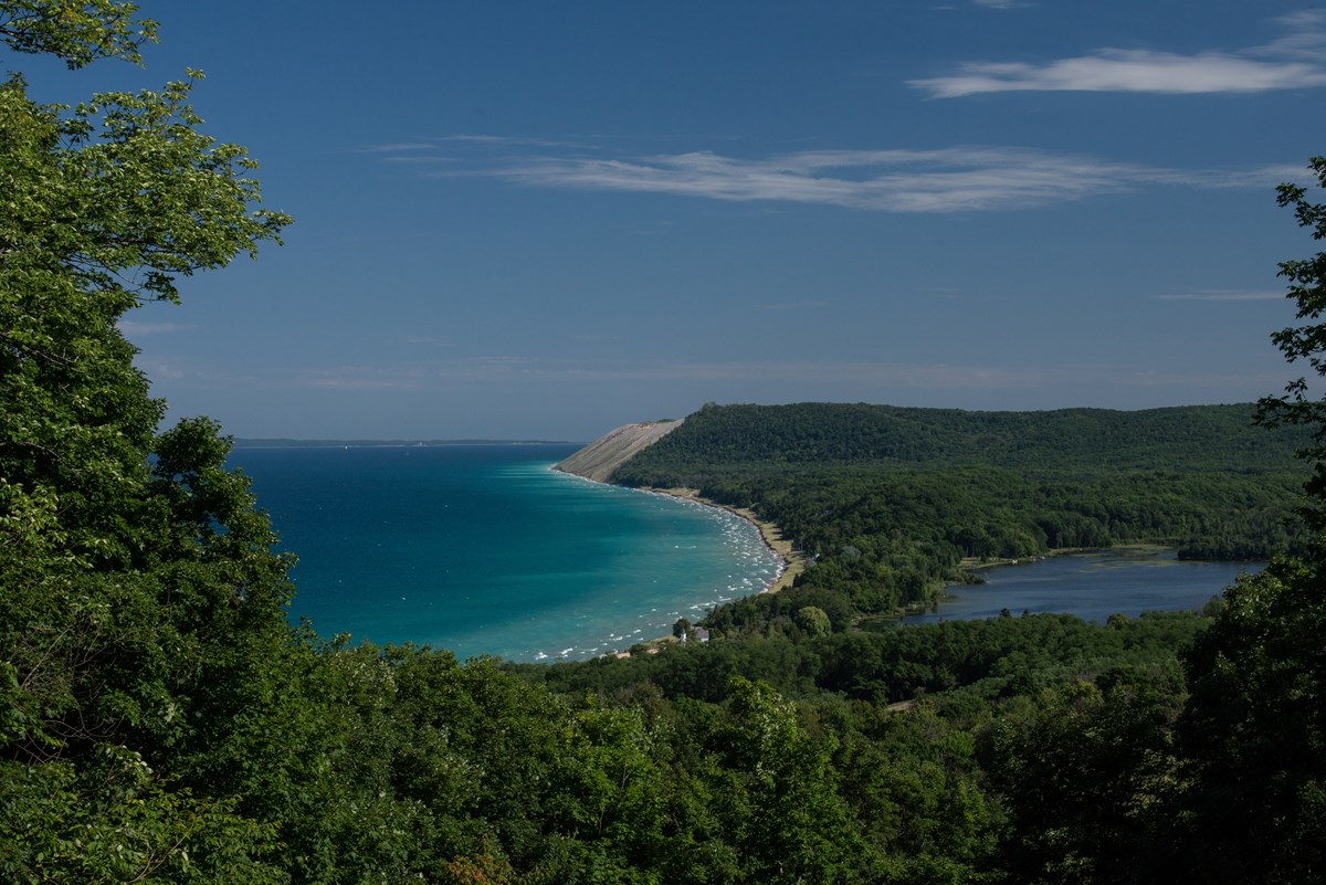 The highpoint view of turquoise water of a lake surrounded by tall sand dunes and lush, green vegetation.