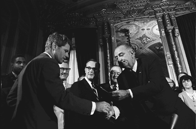 LBJ bends over to hand Bobby Kennedy a pen. Men look on.