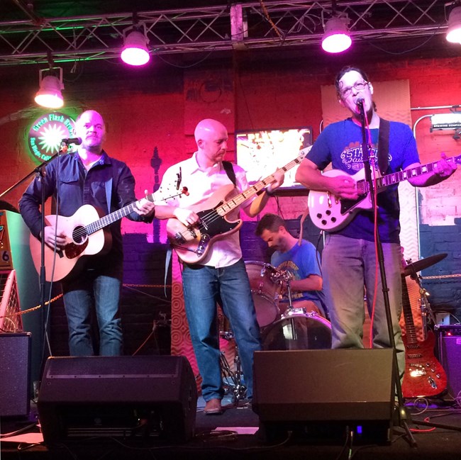 Aaron Lubeck (left) playing acoustic guitar onstage with two bandmates.