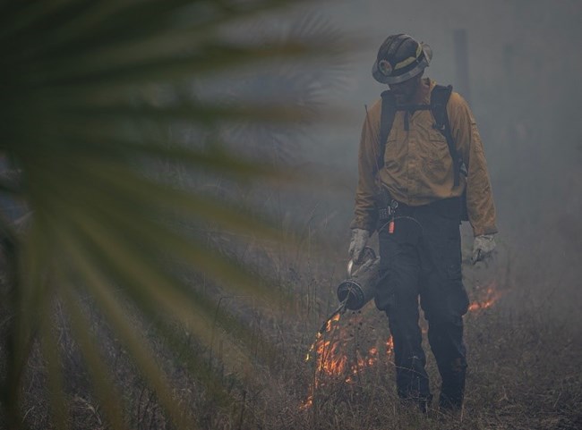 Firefighter uses a drip torch to ignite fire in grass, connecting federal and tribal land.