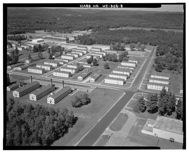 Aerial view of military base with long, white two-story buildings with a long chimney at one end arranged in rows. The buildings are surrounded by grass lawns interrupted by trees, evergreens, and paved roadway