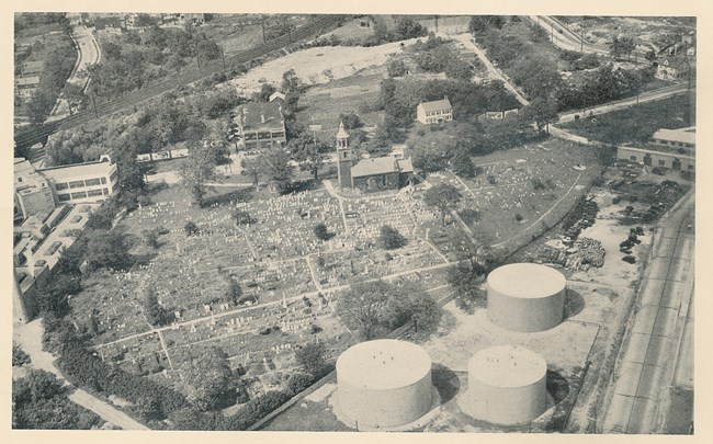Black and white aerial photo, shows cemetery with many gravestones, and adjacent industrial buildings