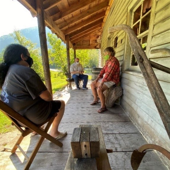 Appalachian Forest NHA Three people wearing masks sit on outdoor heritage furniture on historic wooden porch