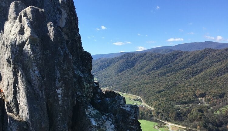 Appalachian Forest NHA View from mountain overlook. Dramatic craggy rock of peak at left. Looking downward there are small buildings in green valley in distance. Hills and blue sky above in background