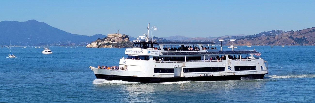 A ferry boat in front of Alcatraz Island. The ferry has visible solar panels and wind turbines, and passengers looking at the water. The side of the boat says "Respect our planet".