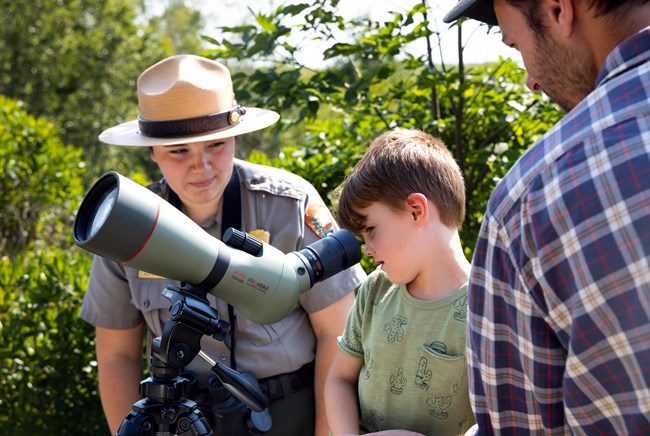 lt= a boy looks through a spotting scope as a park ranger looks on.
