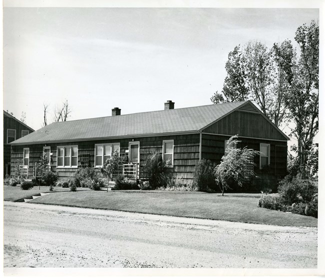 Black & white photo of single-story duplex, ceder shake siding, peaked roof, two doors, four windows