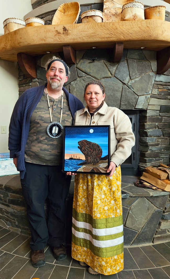 amik received Two people standing in front of a stone fireplace with birch baskets on the mantle. One person wears a yellow ribbon skirt and holds a painting of a beaver.