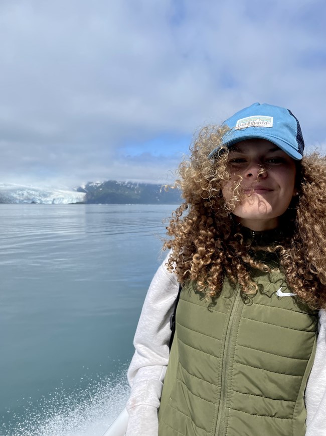 Anna Petr A girl with her hair blowing in the wind with a cap on and glaciers and blue water in the background.
