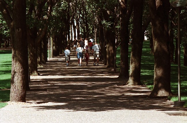 Large, leafy trees line a paved walkway. People are walking along the path.