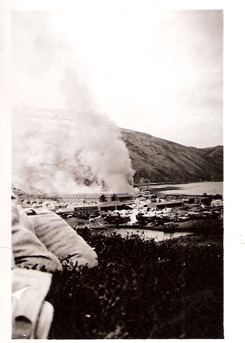 white billowing smoke, water, hills, and buildings below, as seen from on top of a hill, with grass in foreground.