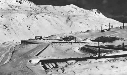 Black and white photo of building in extremely snowy landscape with mountains in background.