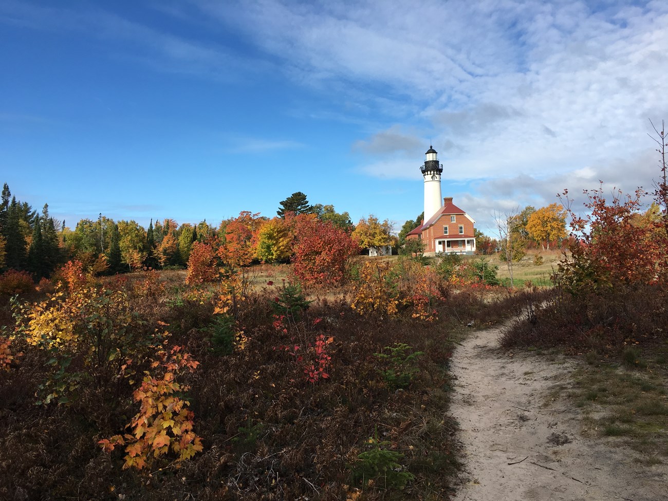 Dirt trail leads to the keepers' quarters and lighthouse in the distance. The leaves on the trees have turned red, orange, and yellow. Dirt trail leads to the keepers' quarters and lighthouse in the distance. The leaves on the trees have turned red, orange, and yellow.