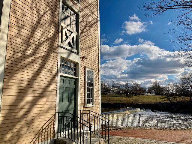 Yellow wood-sided colonial-style mill building facade, brick sidewalk and river to right.