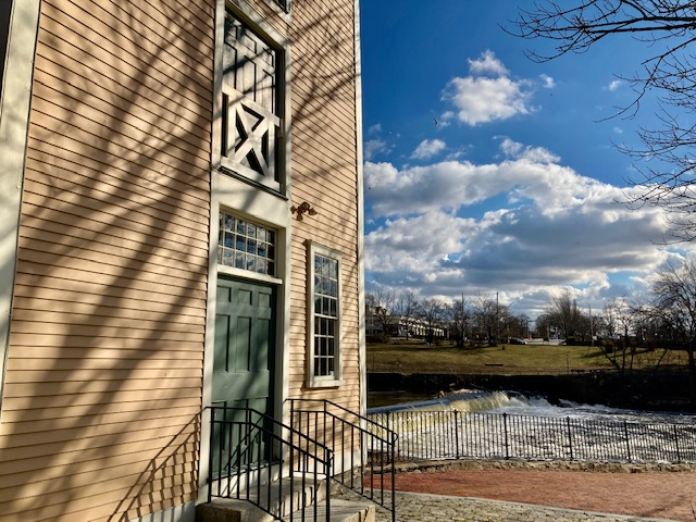 Slater Mill Yellow wood-sided colonial-style mill building facade, brick sidewalk and river to right.