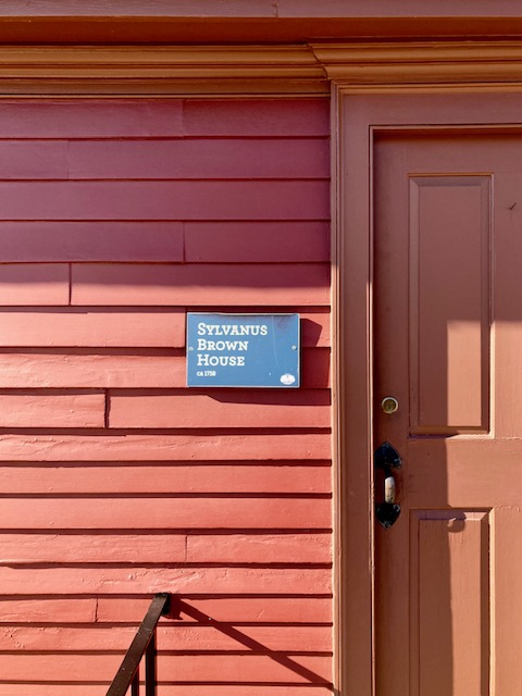 Close-up of facade of red colonial wooden clapboard house with plaque that reads "Sylvanus Brown House, circa 1758