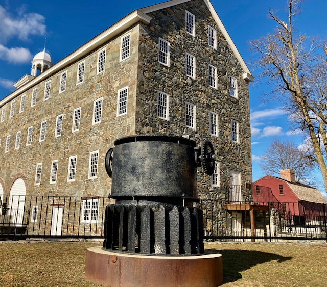 Large grey stone-sided mill with white trim and black metal machinery in front on yard.