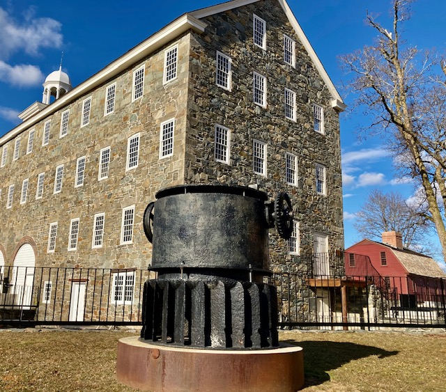 Wilkinson Mill Large grey stone-sided mill with white trim and black metal machinery in front on yard.