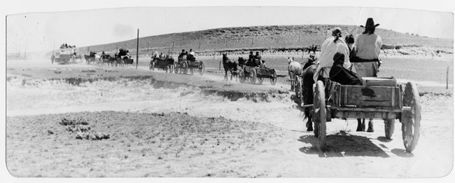 a caravan of horse carriages drives away into dusty prairie in a black and white photo