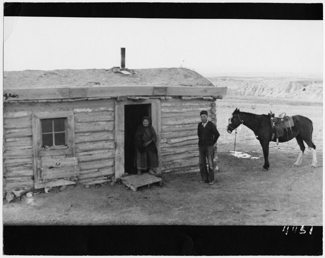 in a historic photo, a man, woman, and horse stand in front of a simple house with badlands formations in the background.