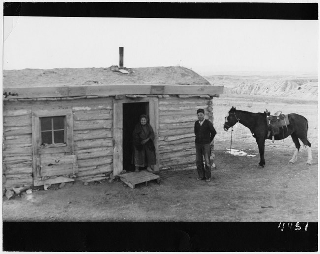 lt= in a historic photo, a man, woman, and horse stand in front of a simple house with badlands formations in the background.